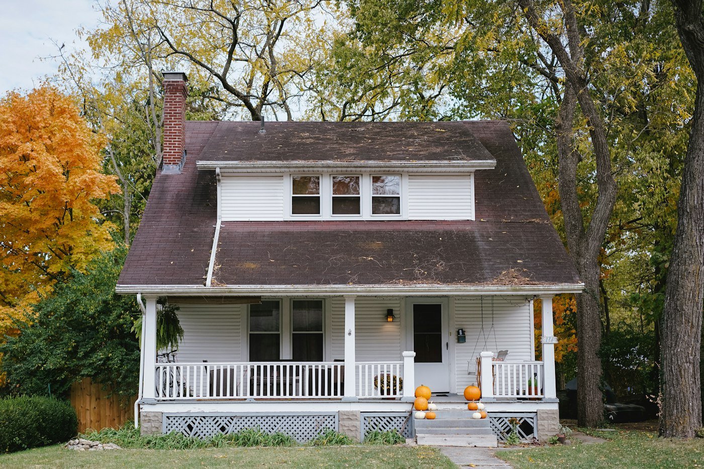 A white house with a dark roof, a front porch, and pumpkins on the steps, surrounded by autumn trees